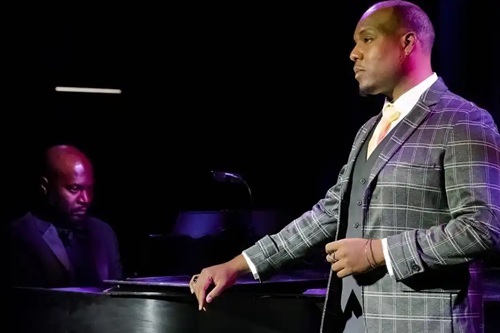 Man singing on stage alongside a piano.